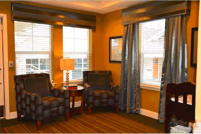 A cozy sitting area with two patterned armchairs separated by a small round wooden table with a lamp and a book on it. The room has mustard yellow walls, striped carpet flooring, and three windows with white blinds and brown valances. There are framed pictures on the walls and a dark wooden piece of furniture partially visible on the right.