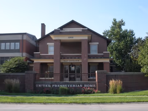 Front exterior view of the United Presbyterian Home building, a brick structure with a main entrance under a covered porch, surrounded by a low brick wall and landscaping including grass, bushes, and trees.