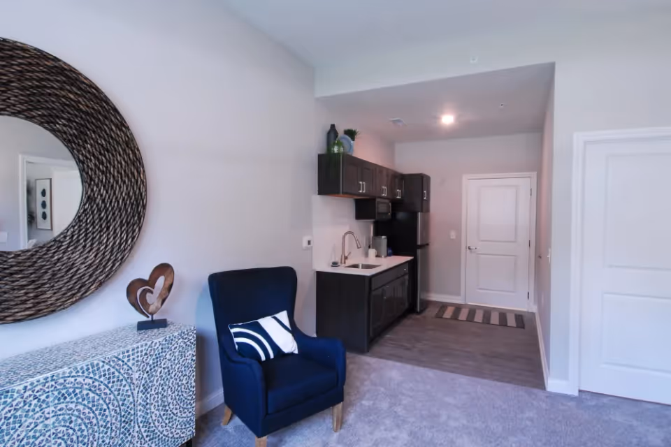 Studio living area with a navy armchair and decorative round mirror beside a small kitchenette with dark cabinets.