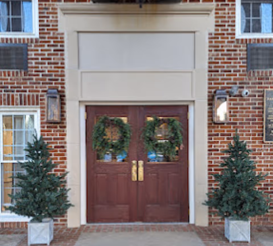 Brick building entrance with double wooden doors and wreaths flanked by potted evergreens.