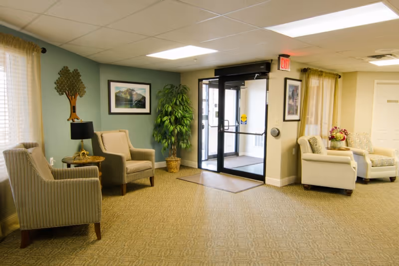 A cozy seating area in a senior living facility with four armchairs arranged around two small tables. The room has a carpeted floor, a potted plant, framed pictures on the walls, and a glass exit door with an illuminated exit sign above it. Soft natural light enters through windows with sheer curtains.