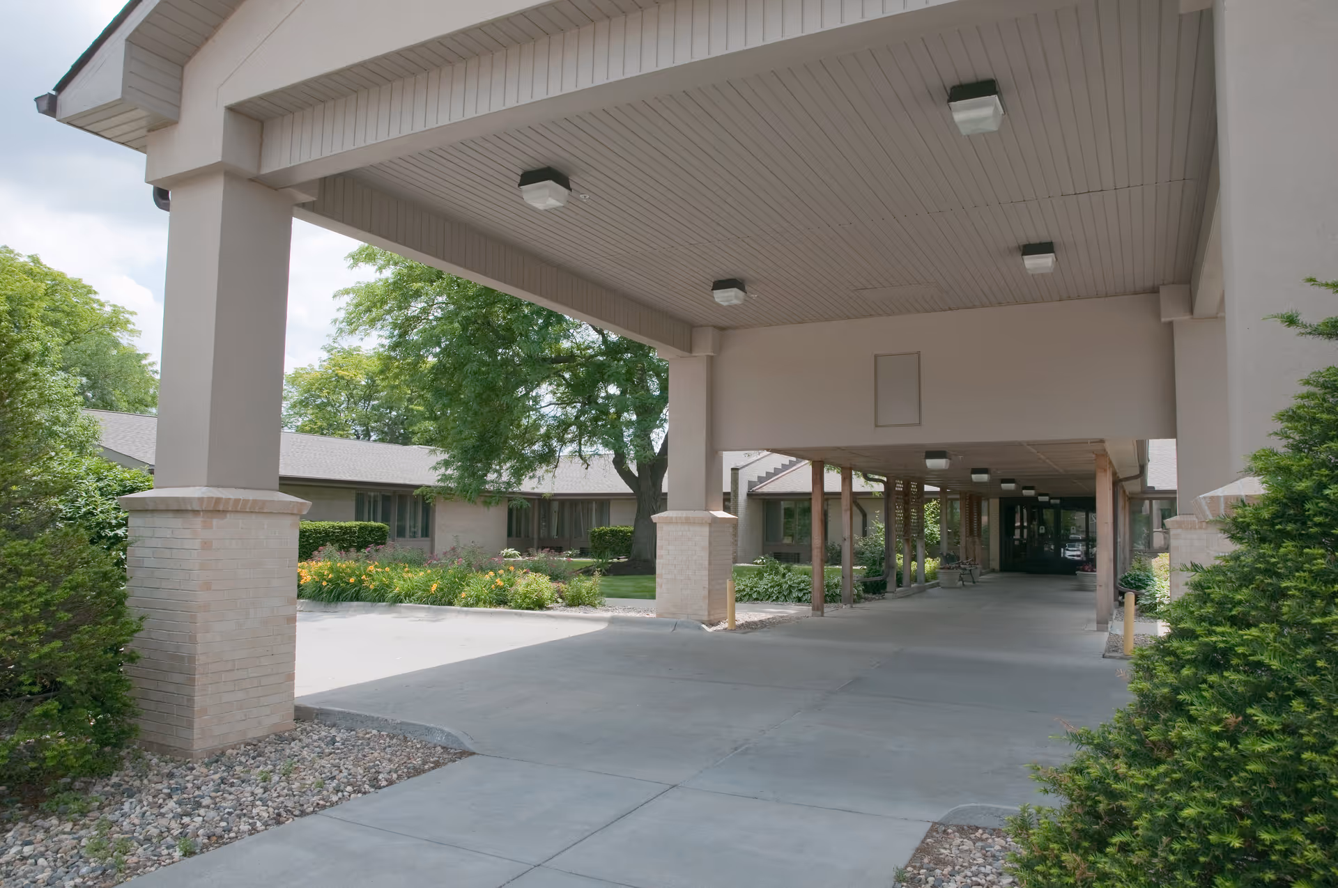 Covered porte-cochere and driveway leading to the front entrance of a single-story senior living building with shrubs and trees.