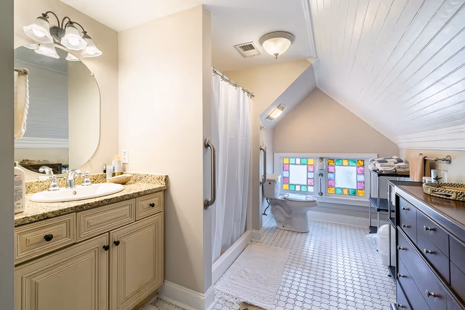 Attic-style bathroom with a vanity and mirror on the left, a shower and toilet near a colorful stained-glass window under a sloped ceiling.