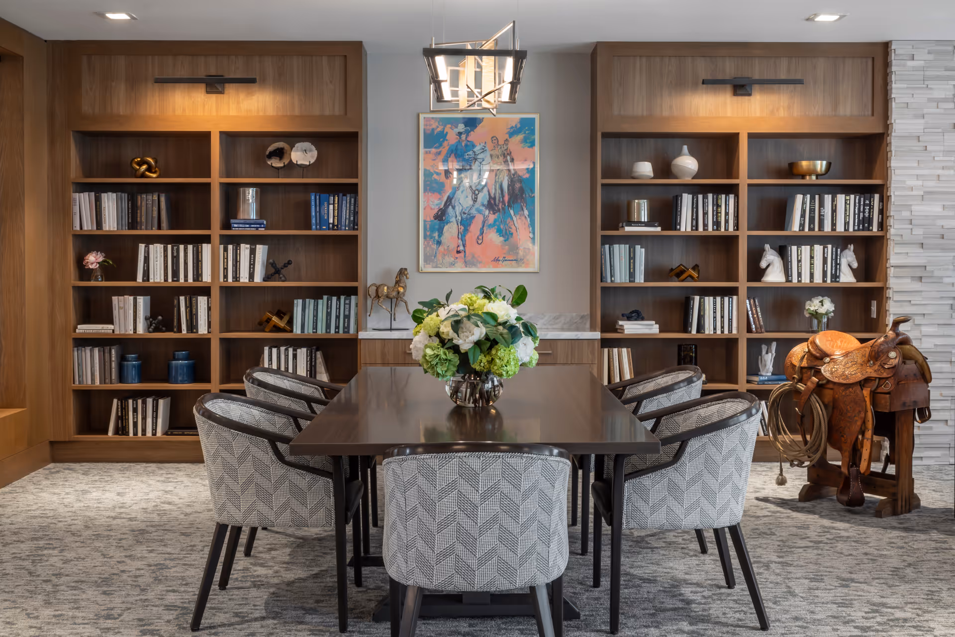 A stylish interior room featuring a rectangular wooden table surrounded by six patterned upholstered chairs. Behind the table are two large wooden bookshelves filled with books and decorative items. A colorful painting of two horseback riders hangs on the wall between the bookshelves. A floral arrangement in a vase is centered on the table. To the right, there is a decorative wooden saddle on a stand. The room has a modern light fixture hanging above the table and a textured white wall on the right side.