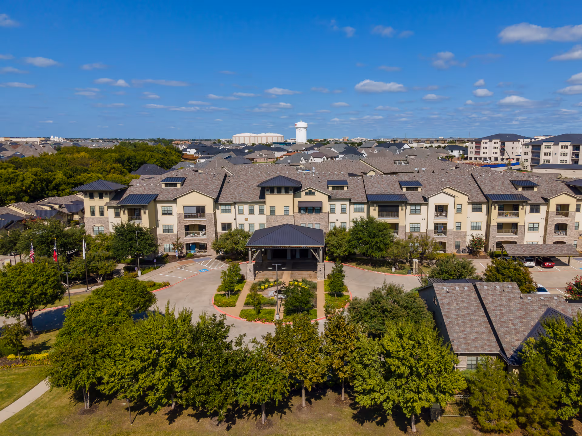 Aerial view of Discovery Village At Twin Creeks, showing a large multi-story residential building surrounded by trees and greenery under a blue sky with scattered clouds. The building has a covered entrance driveway and multiple balconies.