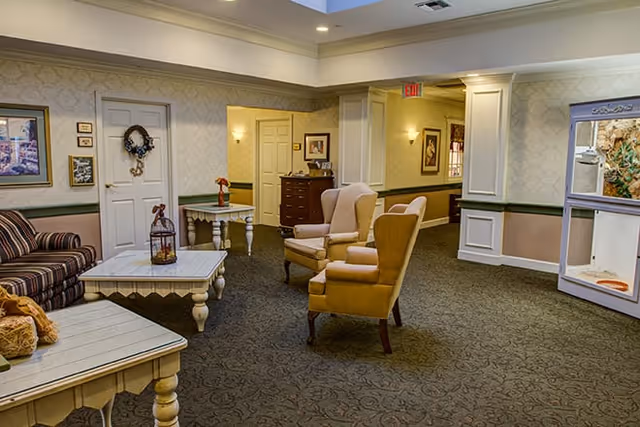 A cozy living room area in a senior living facility with two beige armchairs, a striped sofa, and two white wooden coffee tables. The walls are decorated with framed pictures and a wreath on a door. The carpet is patterned, and the lighting is warm with wall sconces and ceiling lights.