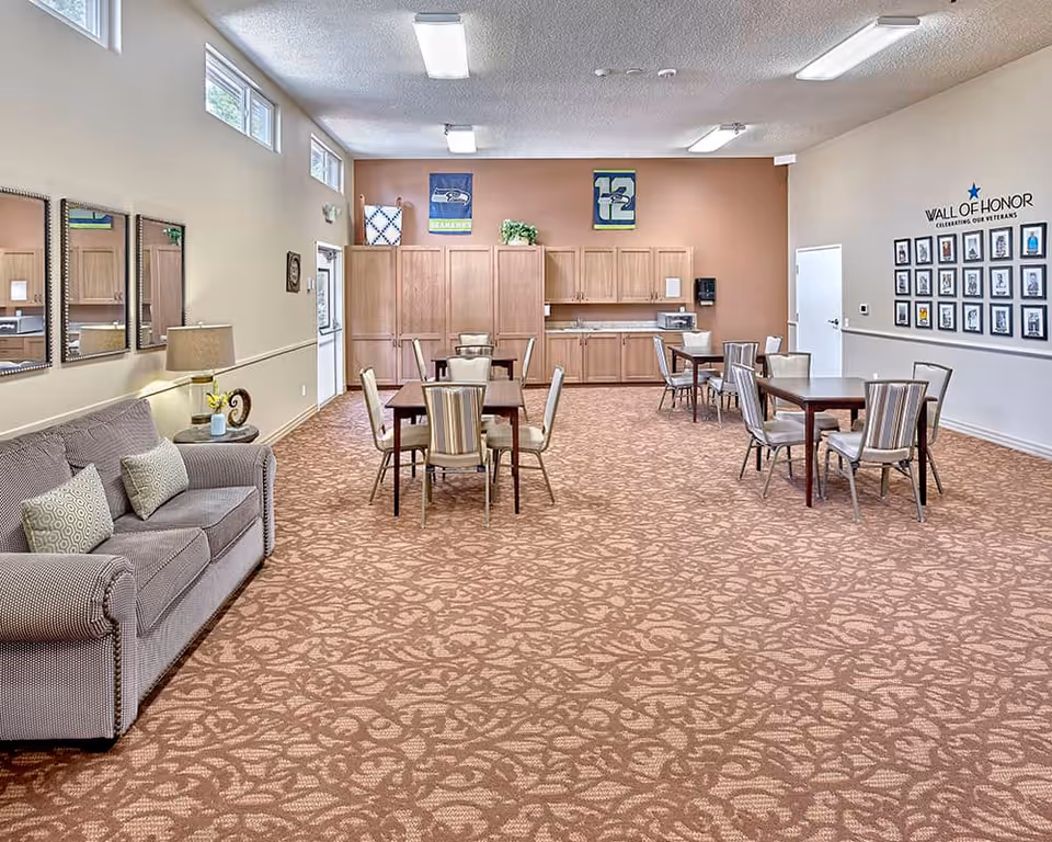 A spacious common area in a senior living facility featuring several tables with chairs arranged for dining or socializing. On the left side, there is a gray couch with patterned pillows and a side table with a lamp and decorative items. The back wall has wooden cabinets, a microwave, and a coffee maker. Above the cabinets, there are two Seattle Seahawks banners. On the right wall, there is a 'Wall of Honor' display with framed photos celebrating veterans. The room has patterned carpet flooring and high windows near the ceiling allowing natural light.