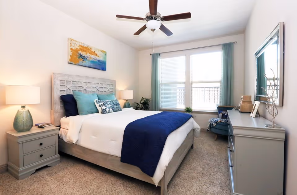 Bright, neatly furnished bedroom with a queen bed, blue accents, bedside tables and lamps, a dresser, and a window with curtains.