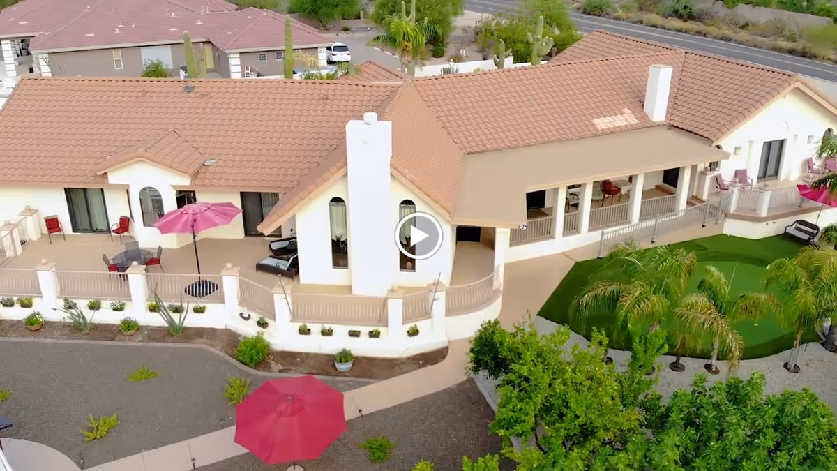 Aerial view of a single-story assisted living building with a tiled roof, wraparound patio featuring pink umbrellas and seating, landscaped grounds and palm trees.