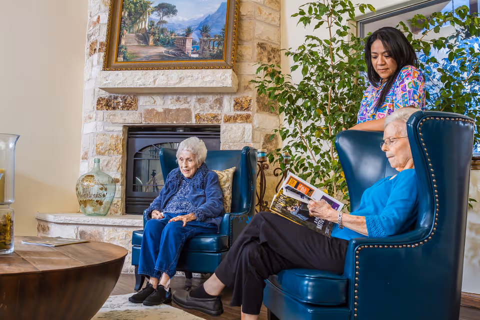 Two elderly women sitting in blue armchairs in a cozy living room with a stone fireplace and a large plant. One woman is reading a magazine while the other is sitting and smiling. A caregiver stands behind them, looking down at the woman reading.
