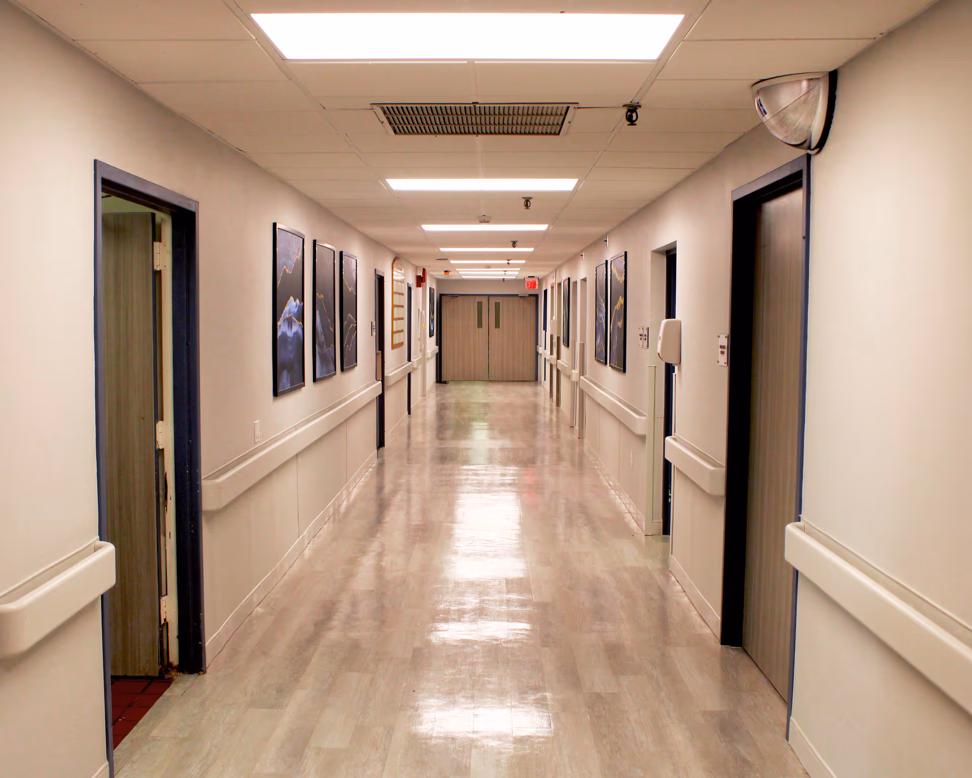A long, clean hallway in a healthcare or senior living facility with closed doors on both sides, handrails along the walls, ceiling lights, and framed artwork hanging on the walls.