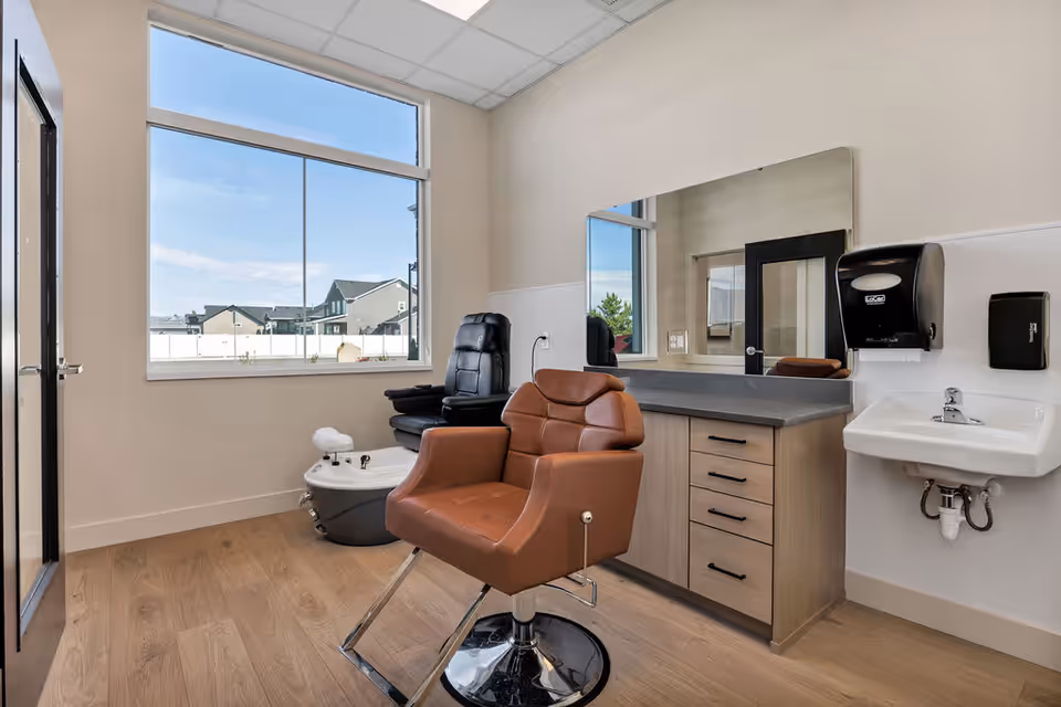 A clean, well-lit interior salon in a senior living facility with a brown barber chair, a sink, and a large window.
