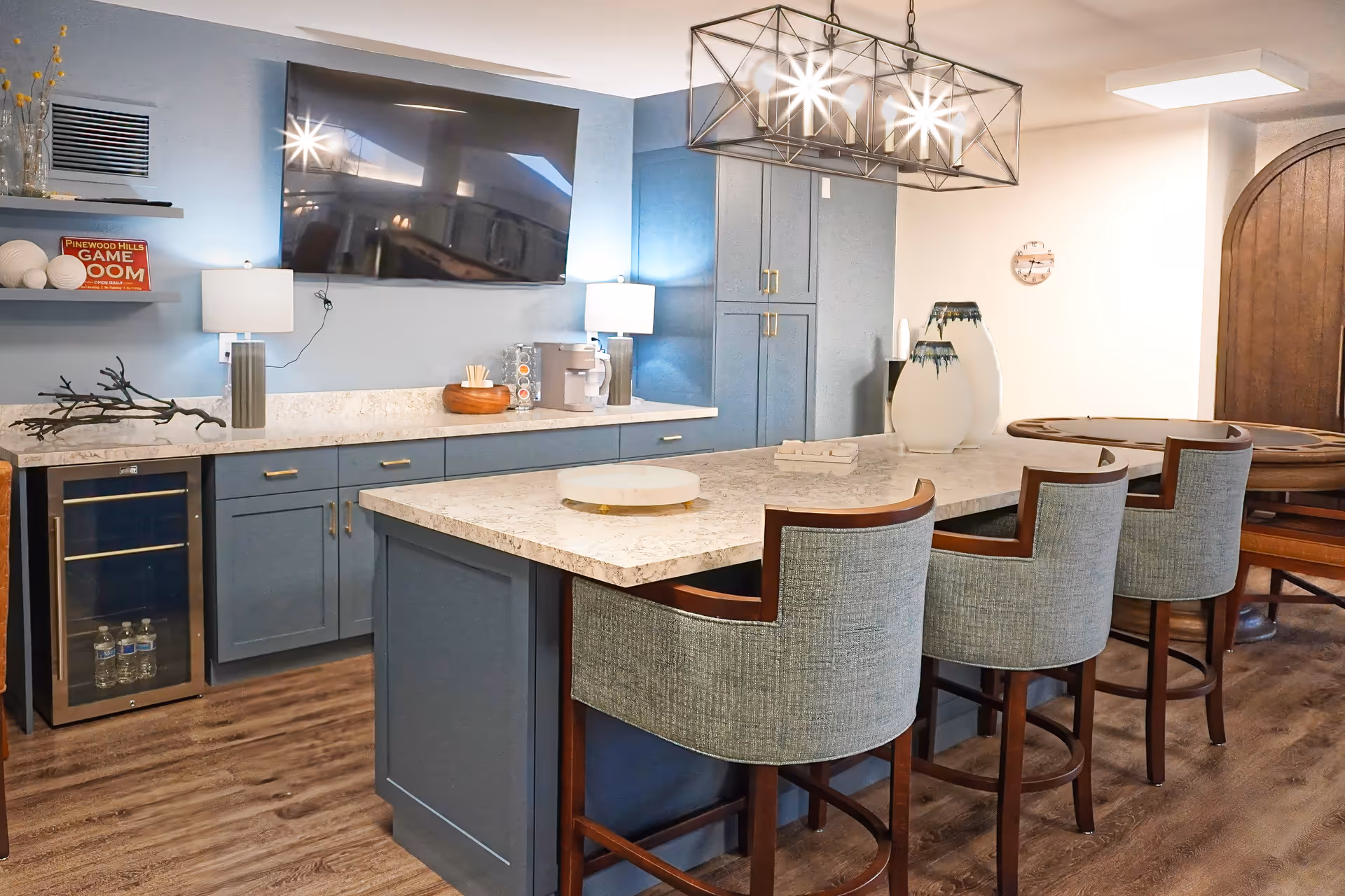 A modern game room with a large flat-screen TV mounted on a blue wall above a countertop with cabinets. There are three upholstered bar stools at a marble island countertop. The room features wood flooring, a wine cooler with bottled water, decorative vases, and a sign that reads 'Pinewood Hills Game Room'. A geometric chandelier hangs above the island, and a round wooden game table is visible in the background.