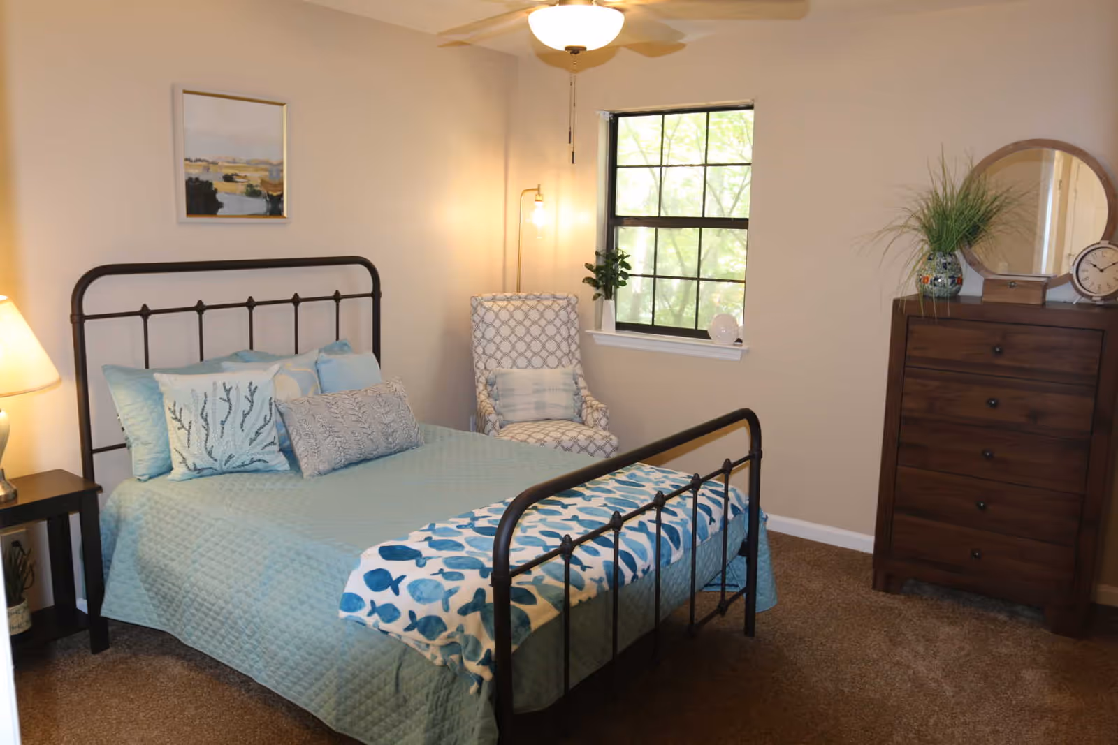 A cozy bedroom with a metal-framed bed dressed in blue bedding, an armchair by a window, and a wooden dresser.