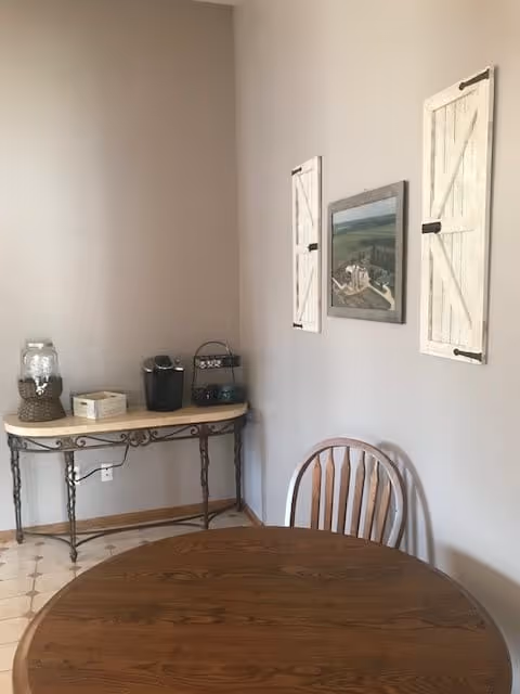 A small dining area with a round wooden table and a wooden chair. Against the wall is a narrow table holding a coffee maker, a glass jar, and a small wooden box. The wall has two white decorative shutters and a framed picture in between them.