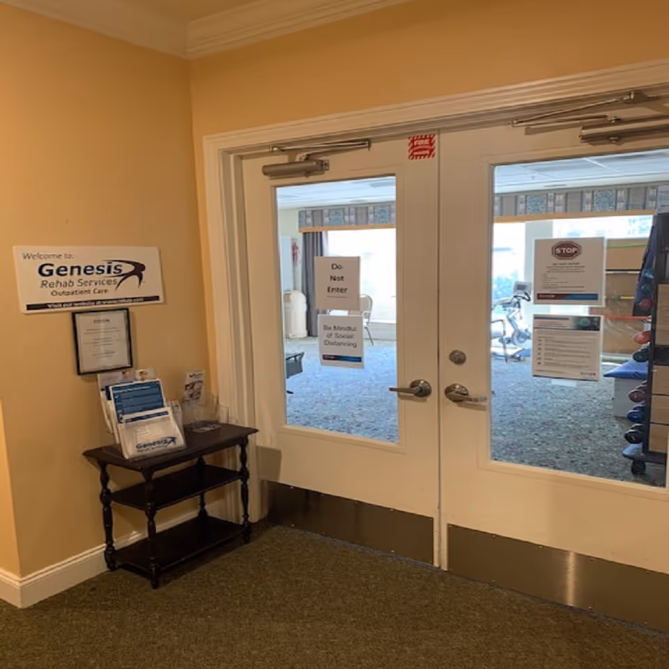 Double glass doors leading into a room with exercise equipment visible inside. A small wooden table with brochures and informational pamphlets is placed against a beige wall to the left of the doors. Signs on the doors read 'Do Not Enter' and 'Be Mindful of Social Distancing'. A sign on the wall says 'Welcome to Genesis Rehab Services Outpatient Care'.