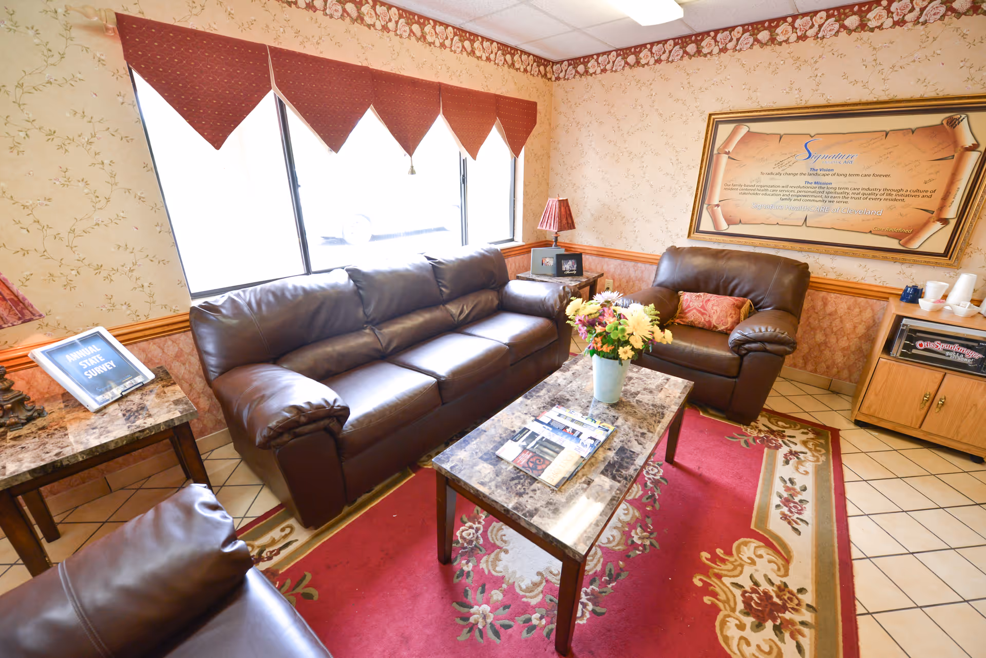 A cozy living room area with brown leather sofas and armchairs arranged around a rectangular marble-top coffee table with a vase of flowers. The room has floral wallpaper, a red patterned rug, and a window with red valances. There is a framed document on the wall, a side table with a lamp and a plaque, and a wooden cabinet with cups and a game box on top.