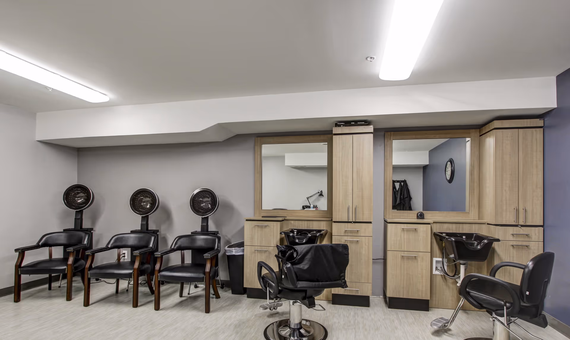 Interior view of a hair salon area with three black hair drying chairs lined up against a gray wall on the left, and two styling stations with mirrors, wooden cabinets, and black salon chairs on the right.