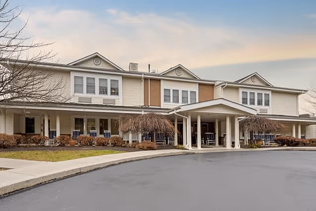 Front exterior of a two-story senior living facility with a covered entrance, porch seating, and a circular driveway.