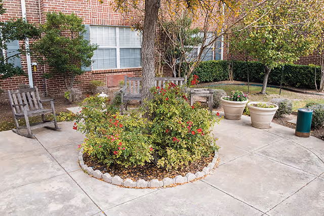 Outdoor courtyard area with a circular flower bed containing a tree and flowering plants, surrounded by a concrete walkway. There are wooden benches and rocking chairs placed around the courtyard, with potted plants and shrubs near a brick building with windows in the background.