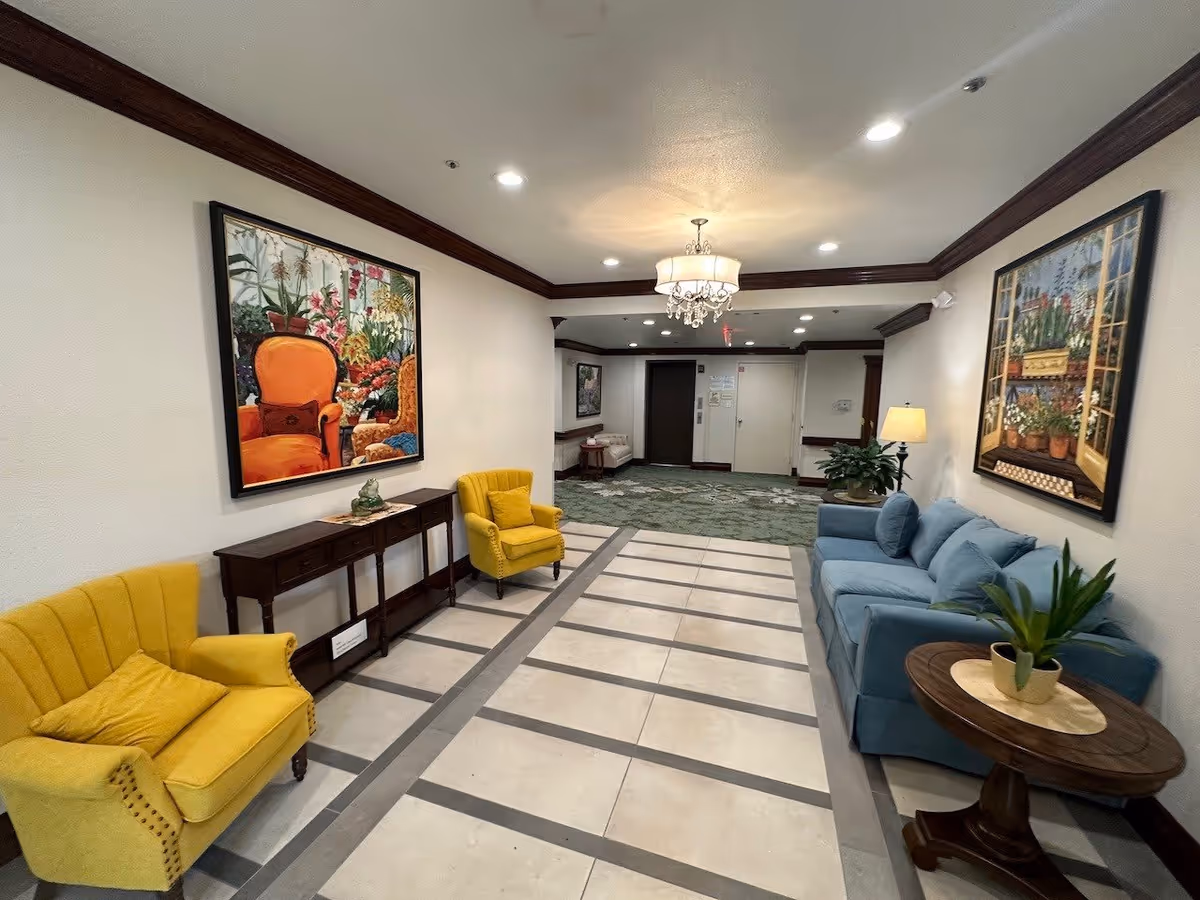 Interior hallway of Tarrytowne Estates featuring a seating area with two yellow armchairs and a blue sofa. The walls are decorated with large colorful paintings of floral and garden scenes. There is a wooden console table between the yellow chairs and a round wooden side table with a potted plant next to the sofa. The floor has a tiled pattern and the ceiling has recessed lighting and a chandelier.