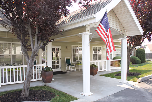 Front entrance of a senior living facility with a covered porch supported by white columns, an American flag hanging, rocking chairs on the porch, potted plants, and trees with red leaves in the landscaped area.