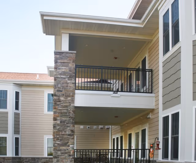 Exterior view of a senior living facility showing a covered balcony with black metal railings and a stone pillar. The building has beige siding with white trim around the windows and roofline.