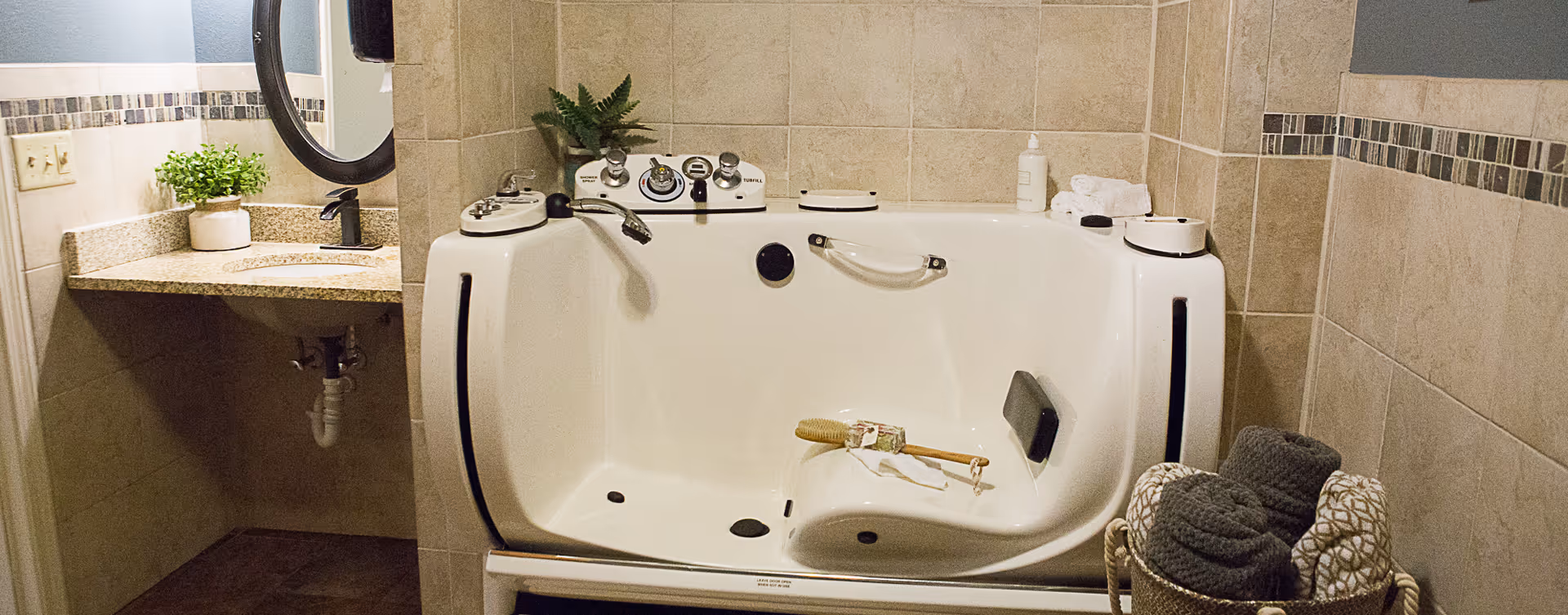 A bathroom featuring a walk-in bathtub with built-in controls and a grab bar. The tub has a brush and soap placed inside. To the left, there is a granite countertop with a sink, a round mirror, and a small potted plant. On the right side of the tub, there is a basket containing rolled towels. The walls are tiled with a decorative border.