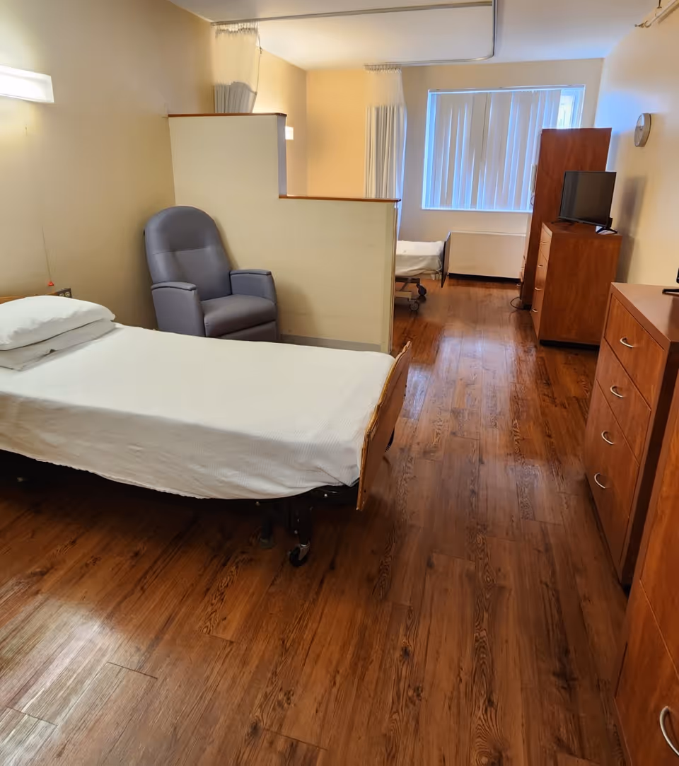 A senior living facility room with two single beds separated by a partial wall with curtains for privacy. The room has wooden flooring, a gray recliner chair, wooden dressers, and a small TV on one dresser. A window with vertical blinds is visible at the far end.