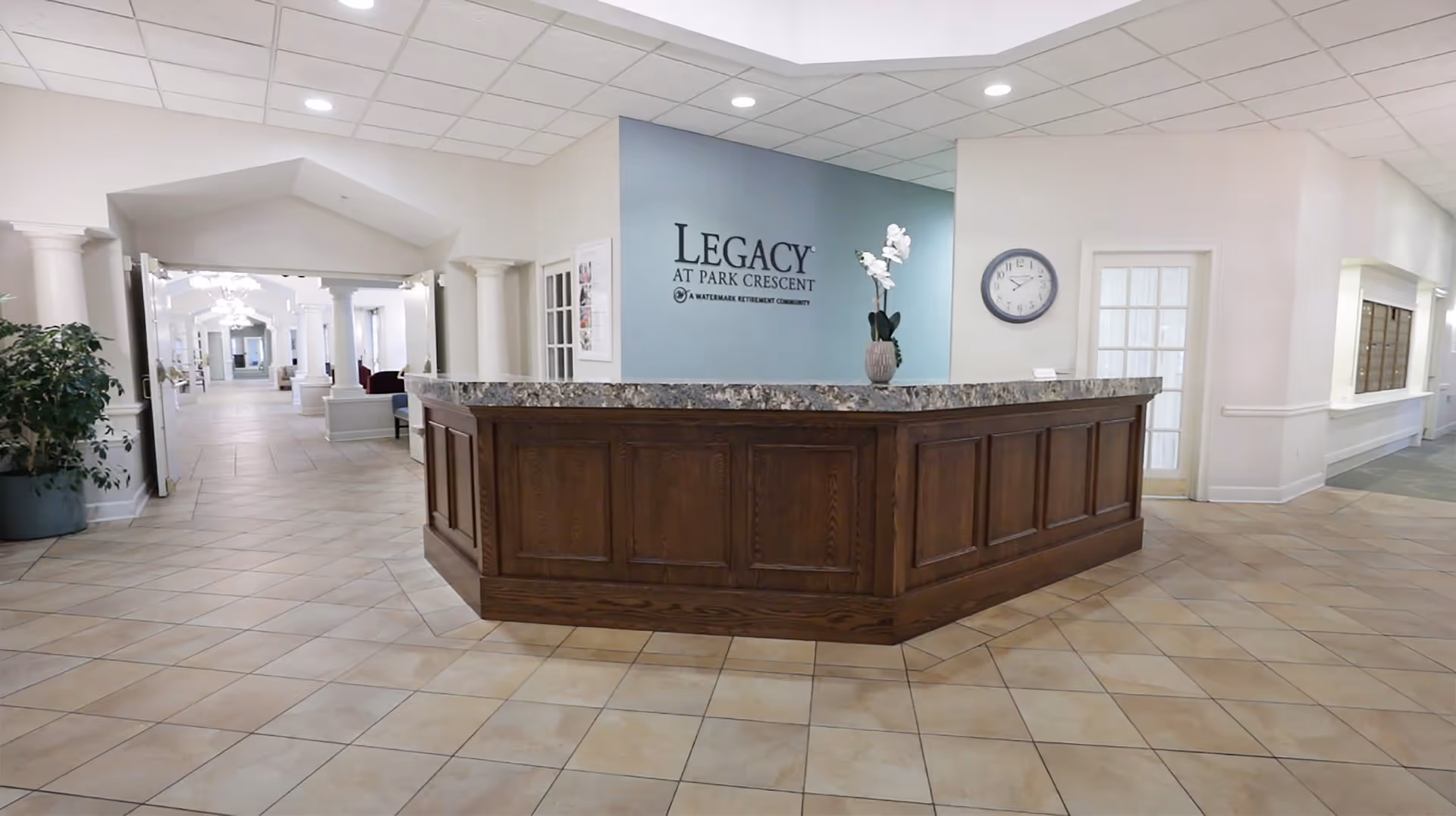 Reception area with a wooden front desk and granite countertop in a senior living facility. Behind the desk is a blue accent wall with the text 'LEGACY AT PARK CRESCENT A WATERMARK RETIREMENT COMMUNITY'. The area has tiled floors, white walls, a clock on the wall, a potted plant, and a hallway with columns and seating visible in the background.