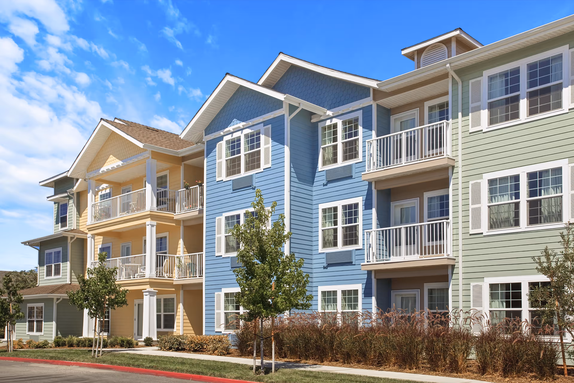 Exterior view of a multi-story residential building with pastel-colored siding in green, yellow, and blue. The building features balconies with white railings, multiple windows, small trees, and landscaping in front under a partly cloudy blue sky.