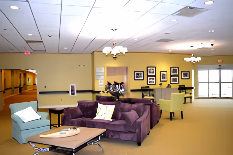 Spacious senior living common lounge with purple sofas, armchairs, tables, and framed pictures on yellow walls.