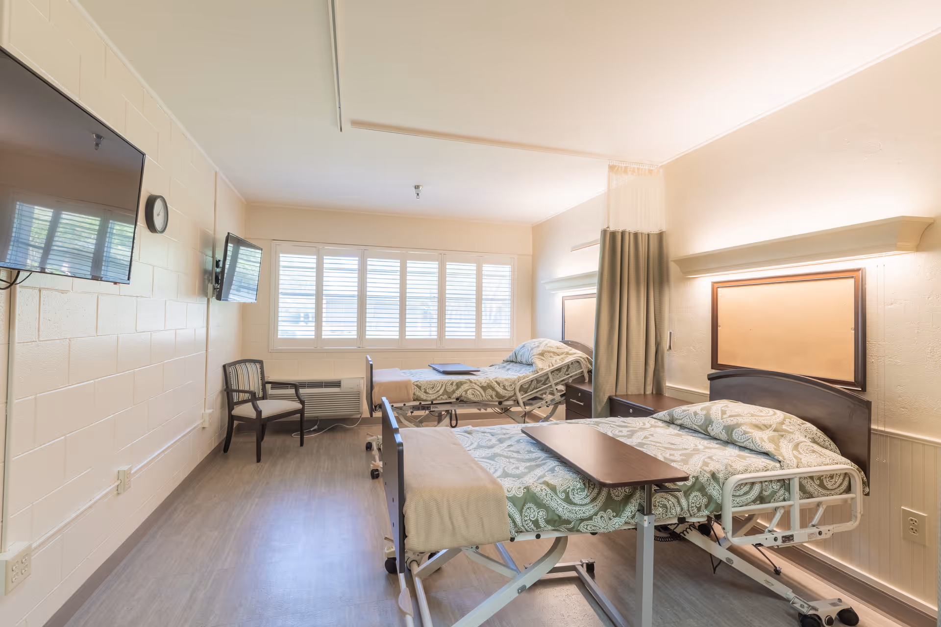 A bright and clean room with two hospital-style beds, each with green patterned bedding and overbed tables. The room has a large window with white blinds, two wall-mounted televisions, a chair, and a privacy curtain between the beds. The walls are light-colored with a clock mounted near one of the TVs.
