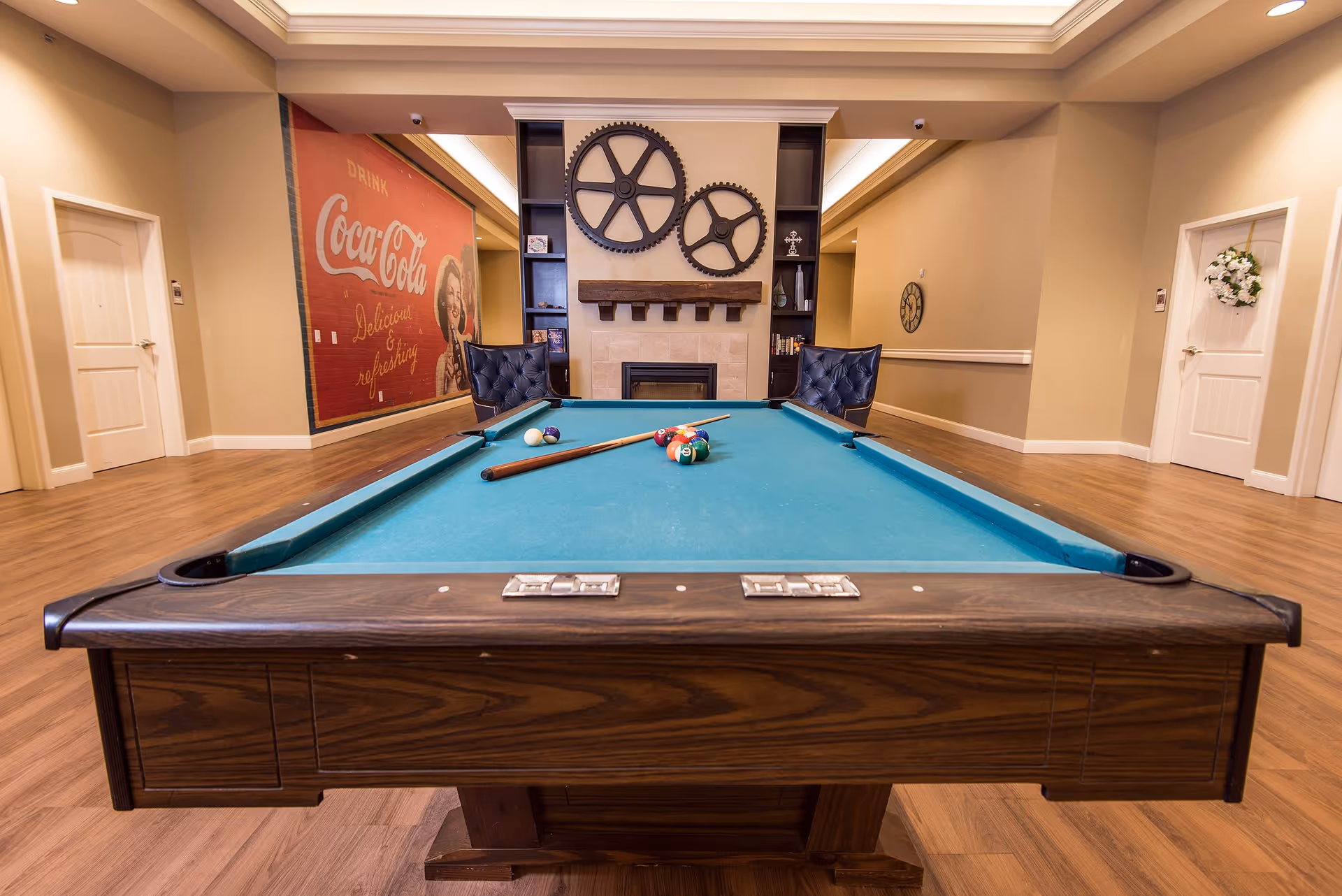 Interior view of a recreational room in a senior living facility featuring a pool table with cues and balls arranged on it. Behind the pool table, there are two black chairs facing a fireplace with decorative large gear wall art above it. On the left wall, there is a vintage Coca-Cola advertisement, and the room has wooden flooring and beige walls with white doors on either side.