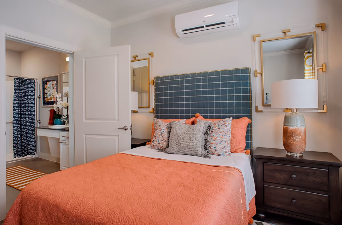 A bedroom with a bed featuring a blue checkered headboard and coral bedding with multiple pillows. There are two matching mirrors on the wall above dark wooden nightstands, one of which has a lamp with a ceramic base. An open door reveals a bathroom with a shower curtain, a vanity with a sink, and decorative items including a vase with flowers.