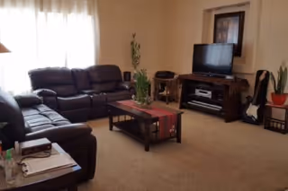 A cozy living room with two dark leather sofas arranged around a wooden coffee table with a red runner and a plant centerpiece. A flat-screen TV sits on a wooden TV stand against the wall, with a framed picture above it. There is a guitar leaning against the wall next to a small table with a potted plant. Light filters in through a window with sheer white curtains.