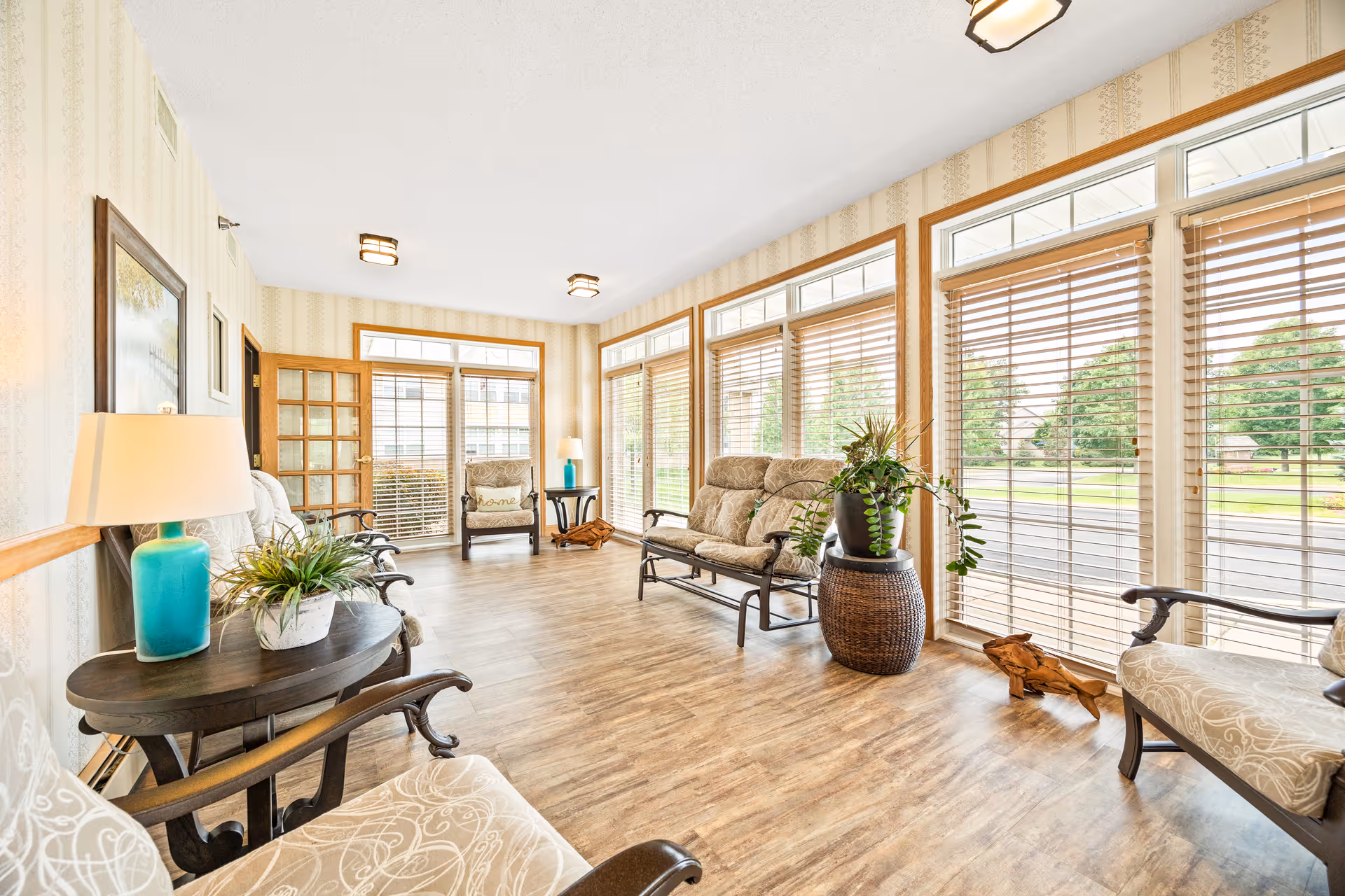Bright sunroom-style sitting area with large windows, wooden floors, cushioned chairs, side tables, lamps, and potted plants.