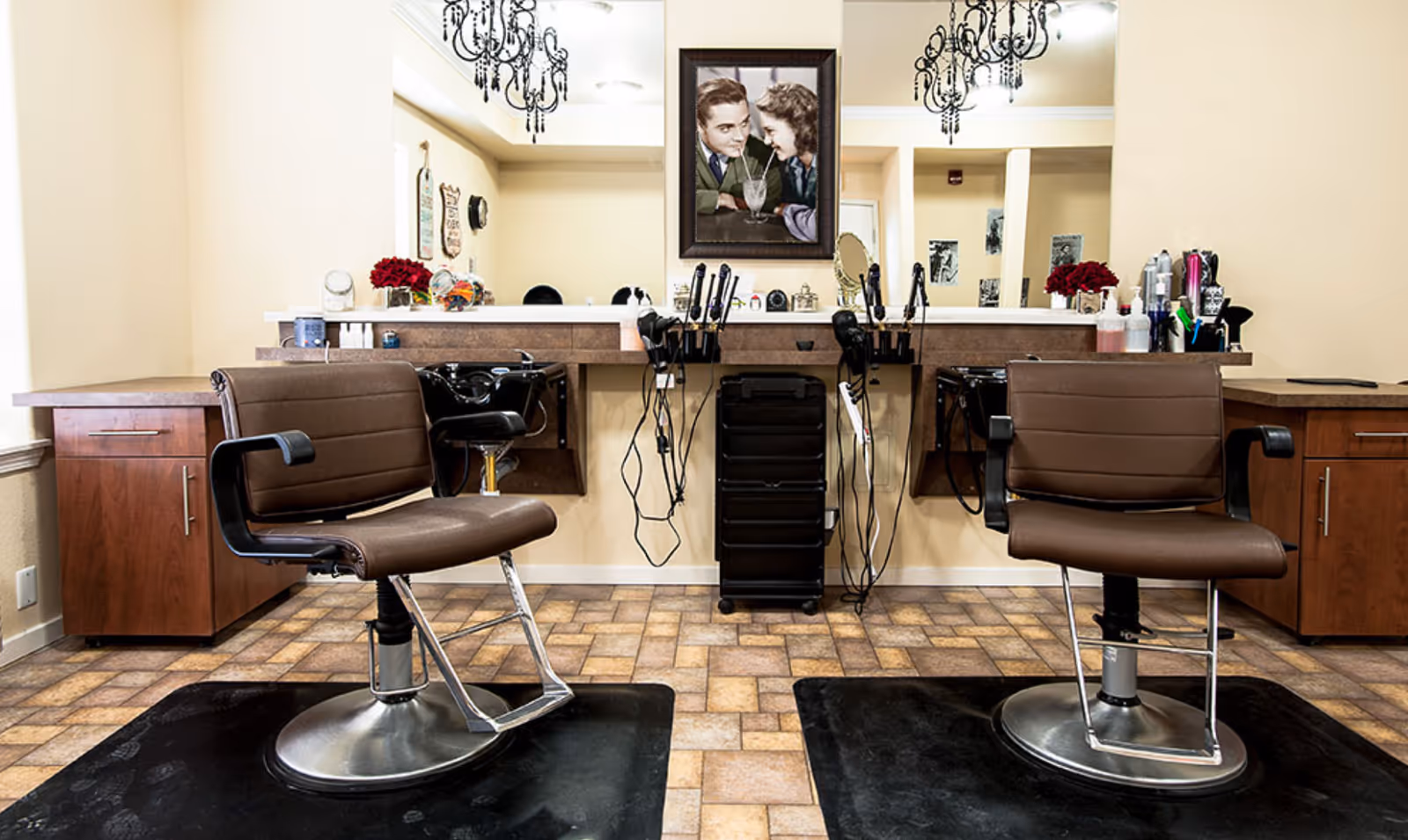Two brown salon chairs facing mirrored styling stations with hair tools, cabinets, and a framed picture on the wall.