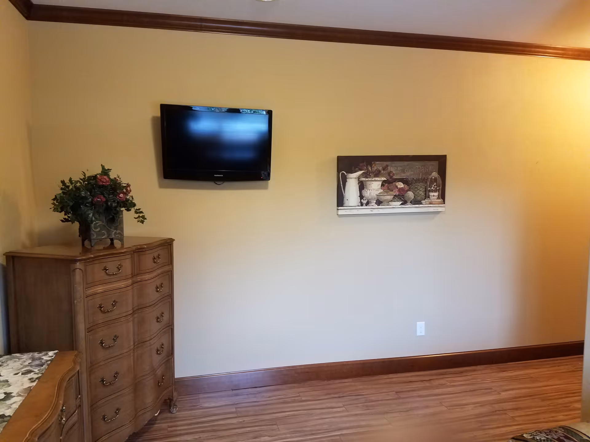 Wall-mounted TV above a wooden dresser with a floral arrangement and a decorative picture on a beige wall in a furnished room.
