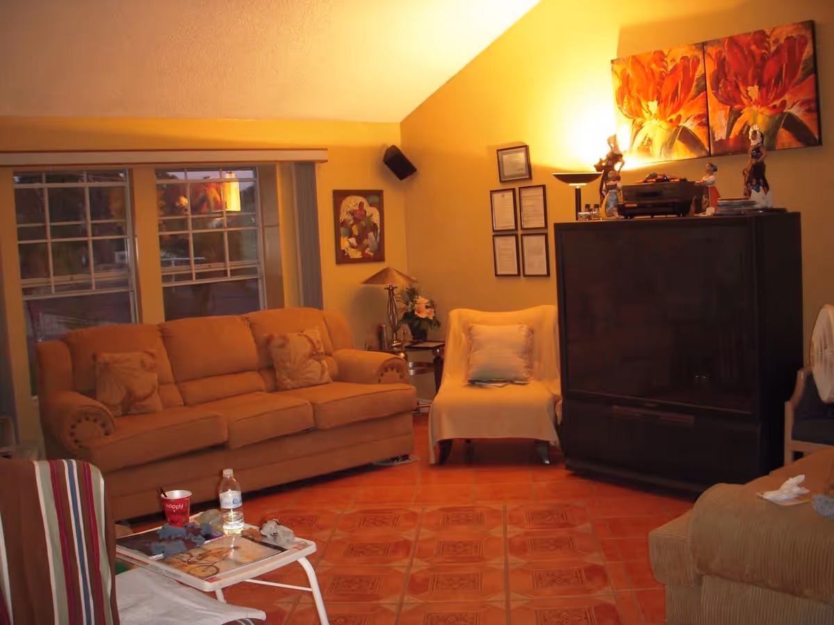 A cozy living room with a brown sofa, a white armchair, a large dark TV cabinet with decorative figurines and paintings above it, a small side table with a lamp and flowers, and a window with vertical blinds. The room has warm yellow walls and tiled floor.