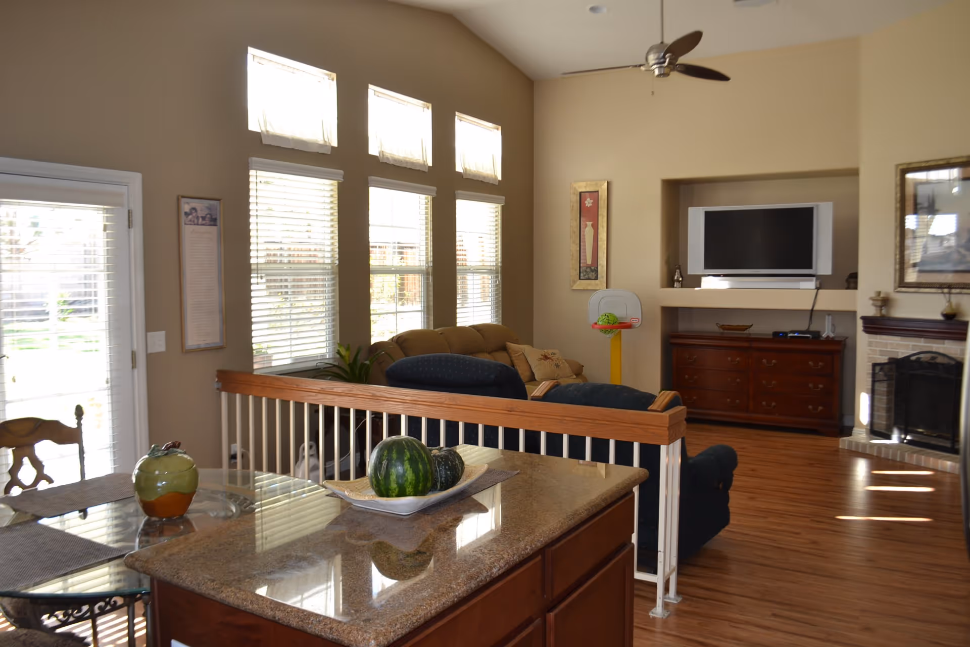 Interior view of a senior living facility common area with a kitchen island in the foreground, a glass dining table with chairs, a living room with sofas, a flat-screen TV mounted above a wooden cabinet, a fireplace, and large windows letting in natural light.