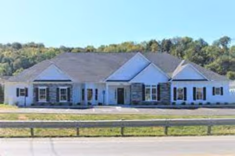 Front exterior view of a single-story senior living facility building with a combination of white siding and stone accents, multiple windows, and a dark shingled roof, set against a backdrop of trees and clear sky.
