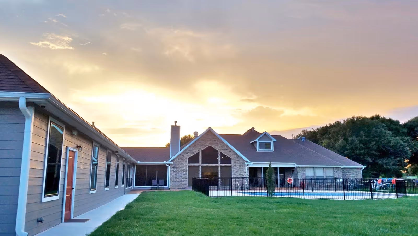 Exterior view of a residential care facility building at sunset with a grassy lawn and a fenced swimming pool area. The building has a combination of brick and siding with multiple windows and a chimney.