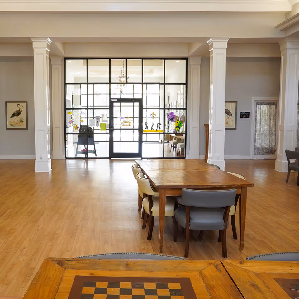 A spacious room with wooden floors and white pillars, featuring a wooden table with chairs around it in the foreground. The background shows a large glass door with black framing, decorated with colorful stickers, leading to a bright area beyond. The walls are light gray with framed bird artwork on either side of the door.