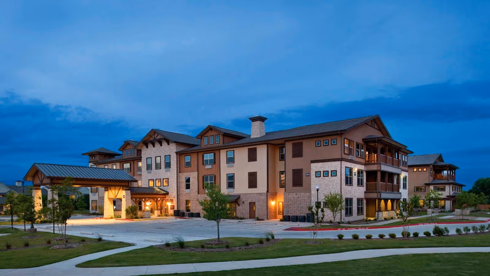 Exterior view of Isle at Cedar Ridge senior living facility at dusk, showing a large multi-story building with stone and wood accents, illuminated entrance, surrounding greenery, and a paved driveway.
