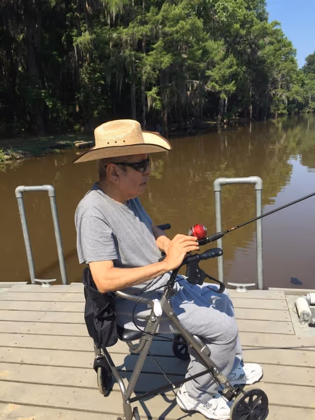 An elderly man wearing a straw hat and sunglasses is sitting on a walker while fishing on a wooden dock by a calm river surrounded by trees.