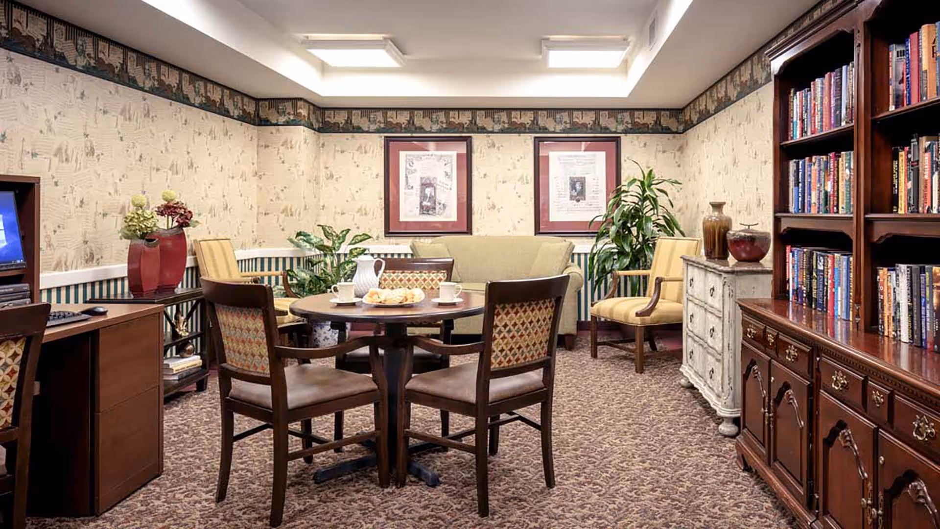 Cozy communal sitting area with a round table and chairs, sofa, armchairs, bookshelves and decorative plants.