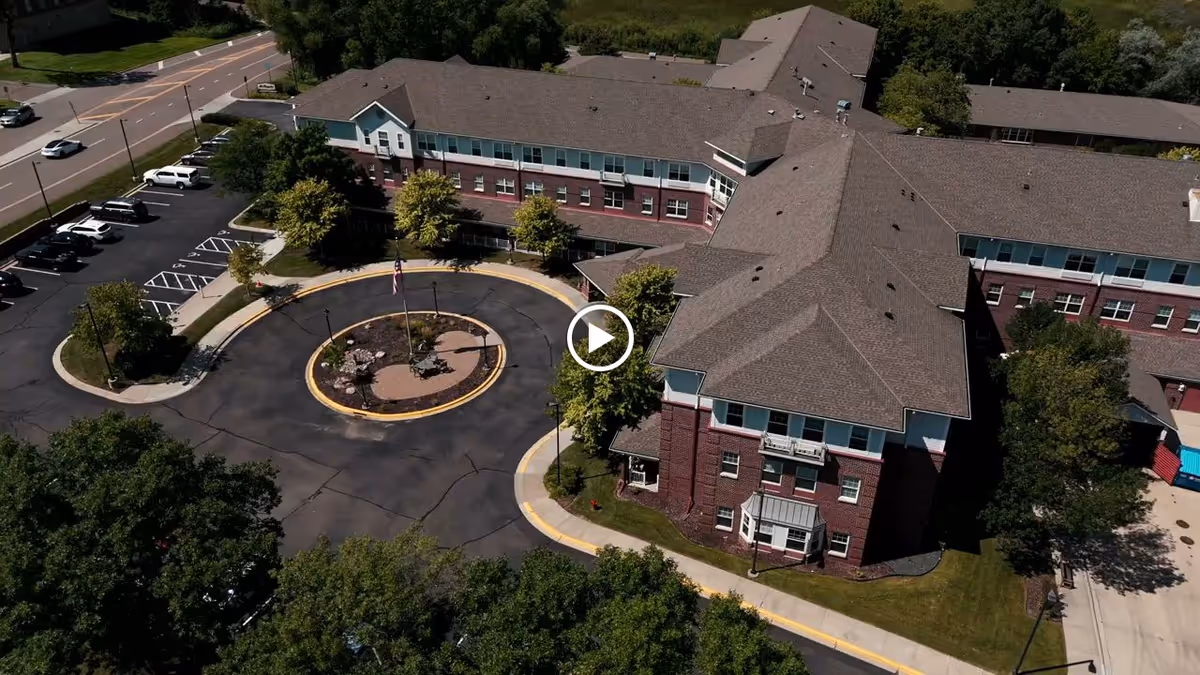 Aerial view of a senior living facility building with a circular driveway and landscaped center island featuring a flagpole. The building is surrounded by trees, parking spaces, and adjacent roads.