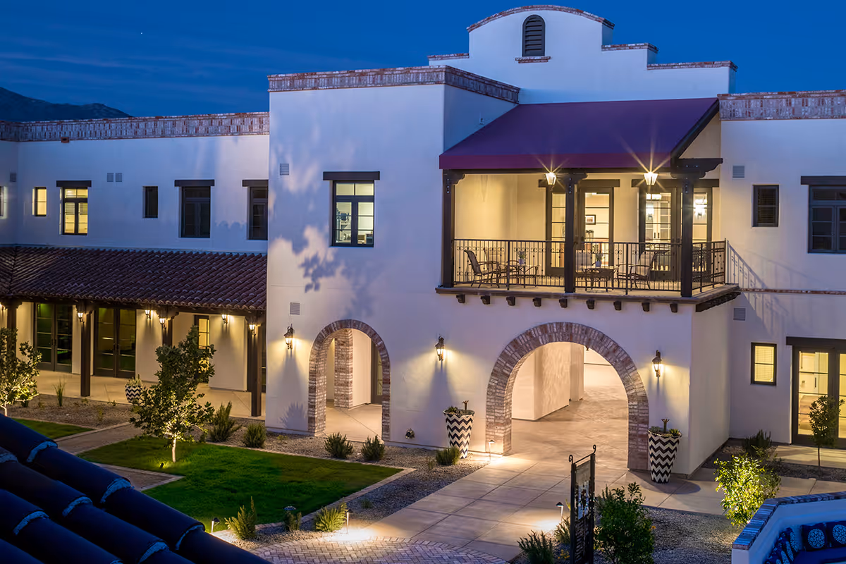 Exterior view of The Hacienda at the River senior living facility at dusk, showing a two-story building with white walls, arched doorways, a balcony with seating under a red awning, and landscaped grounds with small trees and shrubs.