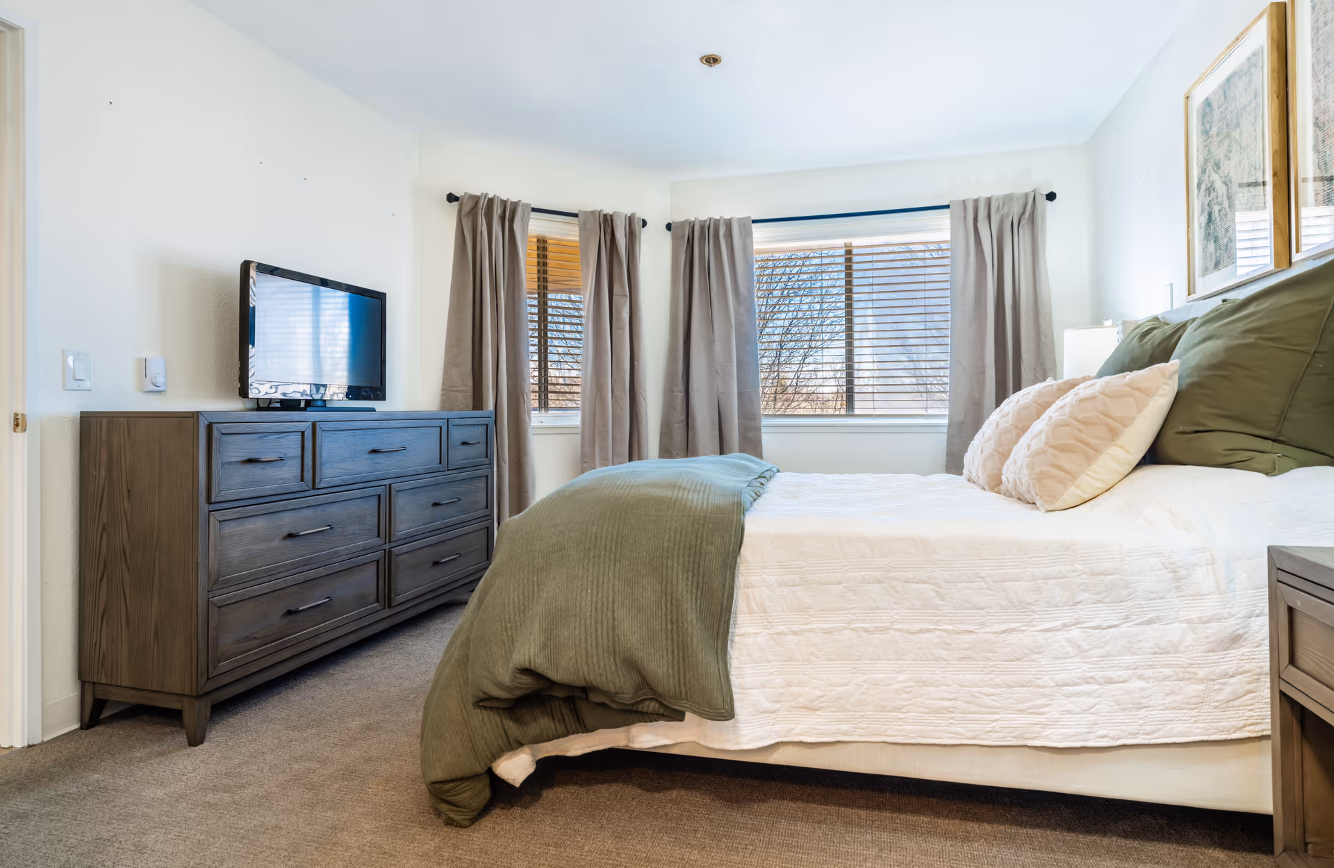 A bright bedroom with a large bed covered in white and green bedding, two framed pictures on the wall above the bed, a wooden dresser with a flat-screen TV on top, and windows with beige curtains allowing natural light to enter.